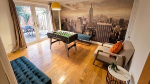 A room with a foosball table, a sofa, a blue ottoman, and a cityscape mural on the wall. - Detached 5-Bed with Games Room · Quedgeley near Gloucester · Sleeps 10 (Quedgeley)