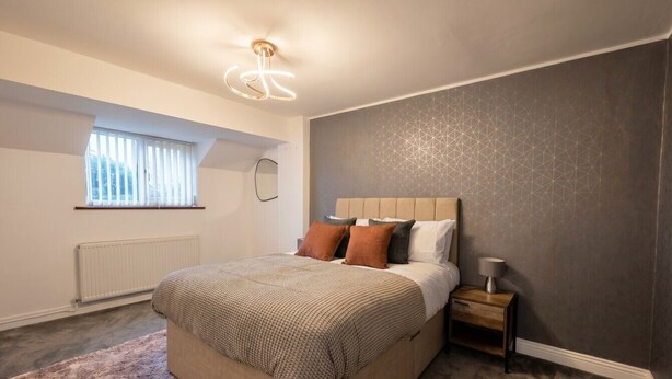 A bedroom with a bed, bedside table, lamp, window, radiator, and a geometric-patterned wall. - Detached 5-Bed with Games Room · Quedgeley near Gloucester · Sleeps 10 (Quedgeley)