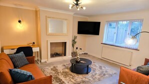 A living room with a sofa, armchair, coffee table, fireplace, TV, and a window with blinds. - Detached 5-Bed with Games Room · Quedgeley near Gloucester · Sleeps 10 (Quedgeley)