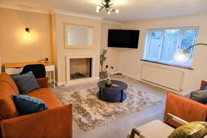 Ein Wohnzimmer mit Sofa, Sesseln, Couchtisch, Kamin, Fernseher und einem Fenster mit Jalousien.