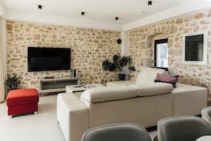 Ein Wohnzimmer mit Steinwand, Flachbildfernseher, beiges Sofa, rotem Hocker und einer Topfpflanze.