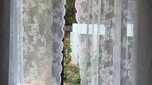A window with white lace curtains, a view of greenery outside, and a glimpse of a building with a green roof. - Tropical Home for Remote Work & Long Stays | Hin Kong (Ko Pha-ngan)