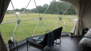 A room with a view of a grassy field through large windows, a bed, a chair, and a small table with a candle and a plant. - Sherwood Pines - Geodome (Mansfield)