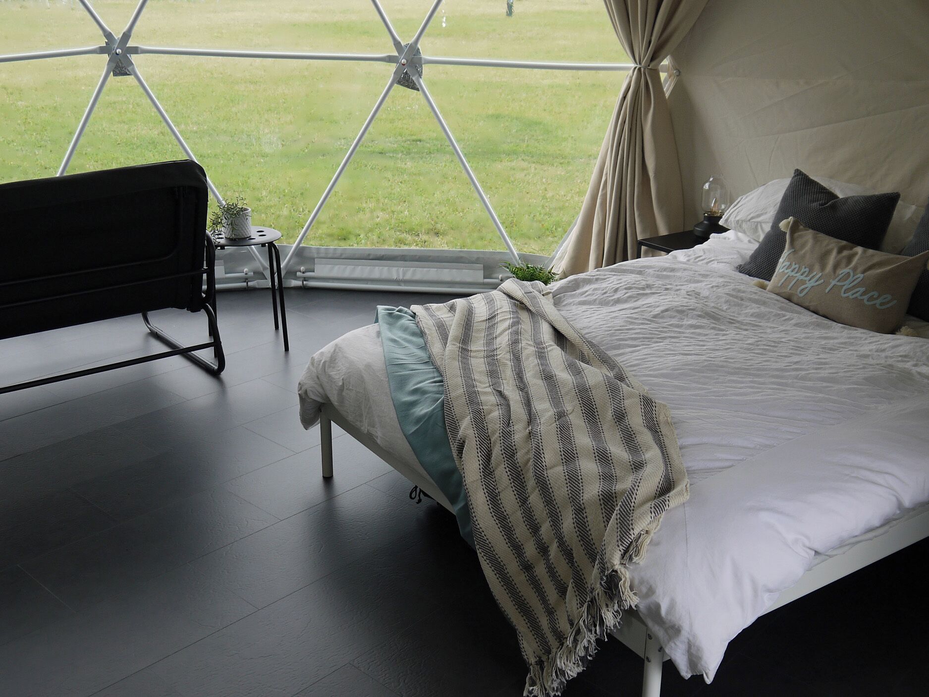 Ein Zelt-Schlafzimmer mit Bett, Kissen, einem Nachttisch und Blick auf ein grünes Feld.