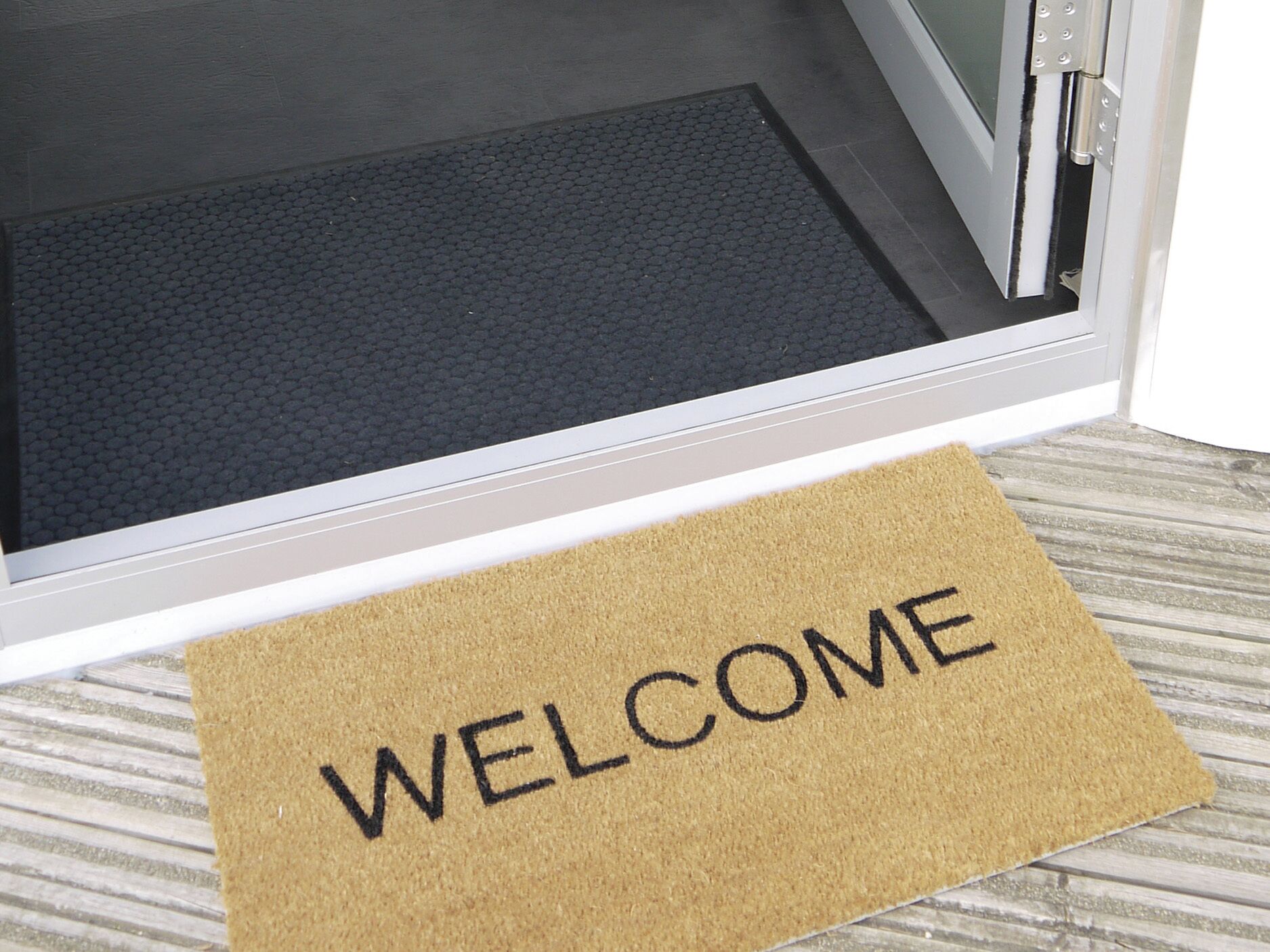 Un felpudo con la palabra 'WELCOME' escrita, colocado frente a una puerta con un tapete texturizado.
