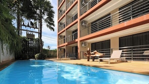 En swimmingpool med liggestole, en flereetages bygning med altaner og palmer.