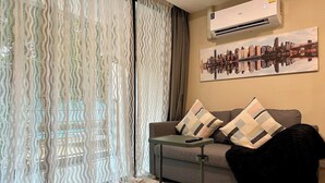 Ein Wohnzimmer mit Sofa, Tisch, Fernbedienung, schwarzer Jacke, Kissen mit Muster, Stadtbildgemälde und einem Fenster mit transparenten Vorhängen.