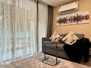 A living room with a sofa, a table, a remote control, a black jacket, a patterned cushion, a cityscape painting, and a window with sheer curtains. - 1BR Apartment Pool Rawai Beach Capital Pro (Rawai)