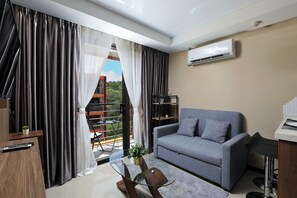 A modern living room with a grey sofa, a glass coffee table, and a view of greenery outside. - 1BR Apartment in Nai Harn Condo | CapitalPro (Rawai)