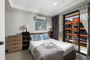 A modern hotel room with a bed, a desk, a chair, a window with curtains, and a view of an orange building. - 1BR Apartment in Nai Harn Condo | CapitalPro (Rawai)