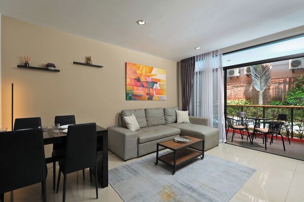 A modern living room with a sofa, dining table, and chairs, a coffee table, and a large window with a view of a balcony. - Studio Pool Access Nai Harn Beach Capital Pro (Rawai)