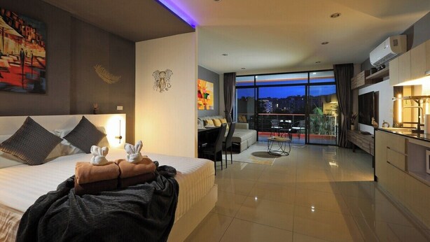 A modern hotel room with a large bed, a desk, and a view of the city at dusk. - Spacious Studio Naiharn Beach Condo | CapitalPro (Rawai)