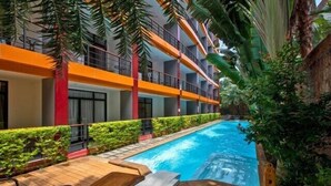 A pool area with wooden loungers, a clear blue pool, and a multi-story building with balconies. - Spacious Studio Naiharn Beach Condo | CapitalPro (Rawai)
