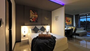 A modern hotel room with a large bed, bedside lamps, artwork on the walls, a dining area, and a view of the outdoors. - Spacious Studio Naiharn Beach Condo | CapitalPro (Rawai)