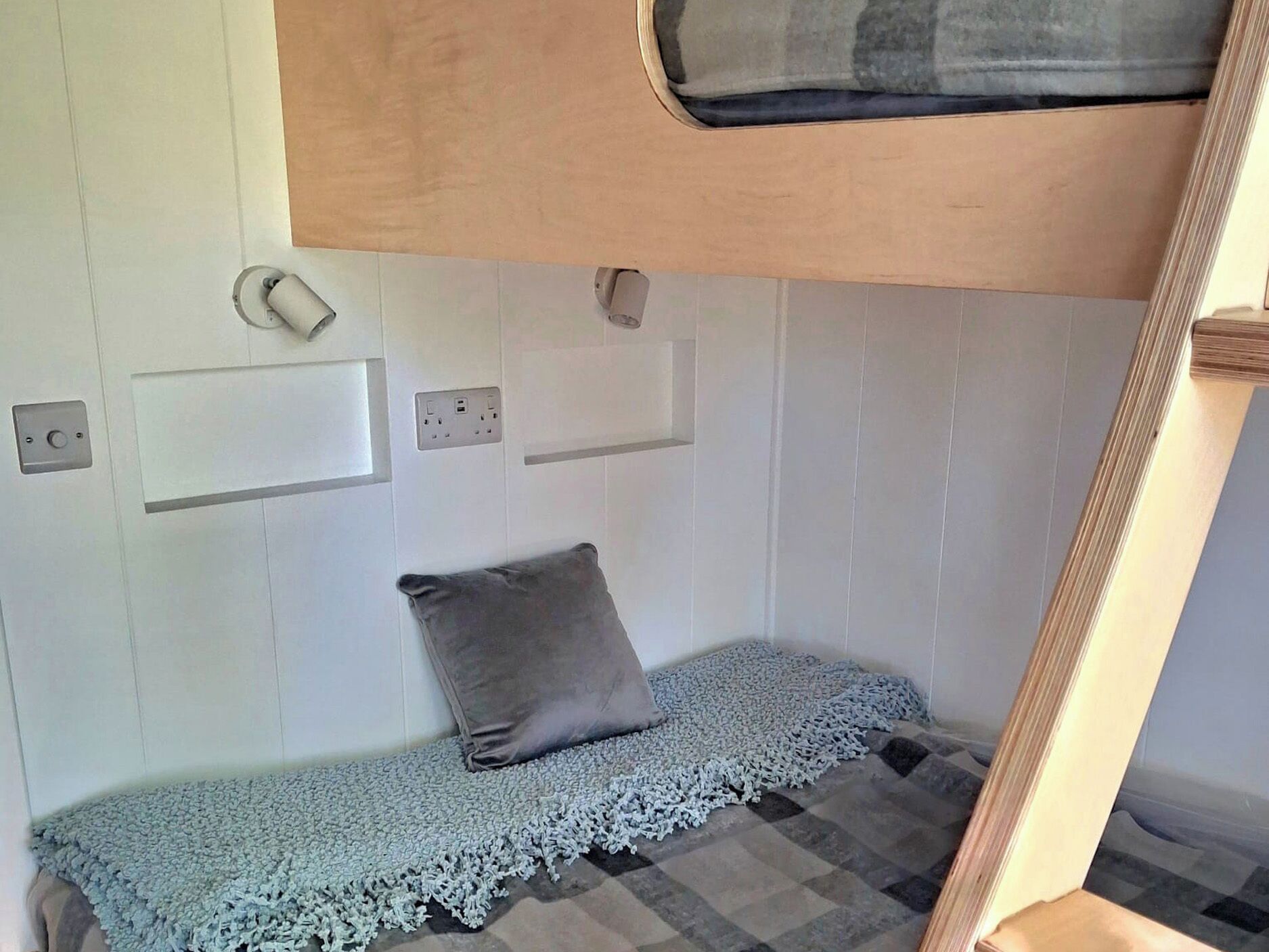 A compact RV interior with a bunk bed, a small seating area with a cushion, and a window.