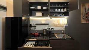 A compact kitchen with a black refrigerator, a stove, a sink, and shelves above the counter holding dishes and glasses. - 1BR Mountain View Nai Harn Beach | Capital Pro (Rawai)