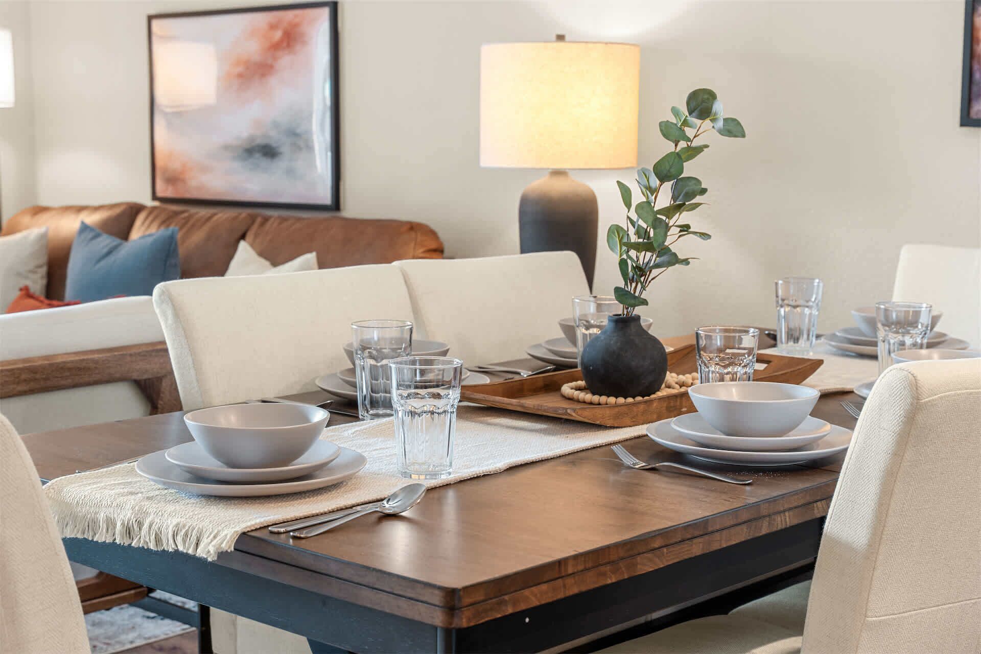 Un coin repas avec une table dressée pour quatre personnes, un vase agrémenté de verdure et une lampe en arrière-plan.