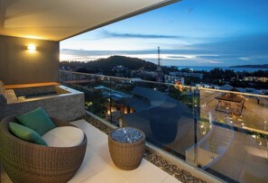 Ein Balkon mit Rattan-Sofa, einem runden Rattan-Hocker und einem Glasgeländer mit Blick auf eine Stadtlandschaft in der Dämmerung.