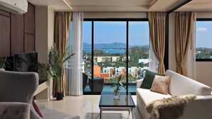 Ein modernes Wohnzimmer mit Sofa, Sessel und Couchtisch. Ein Fernseher auf einem Schrank, eine Topfpflanze und ein Blick auf Stadt und Berge durch große Fenster.