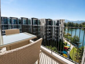 Ein Balkon mit Korbmöbeln, Blick auf einen See und moderne Gebäude im Hintergrund.