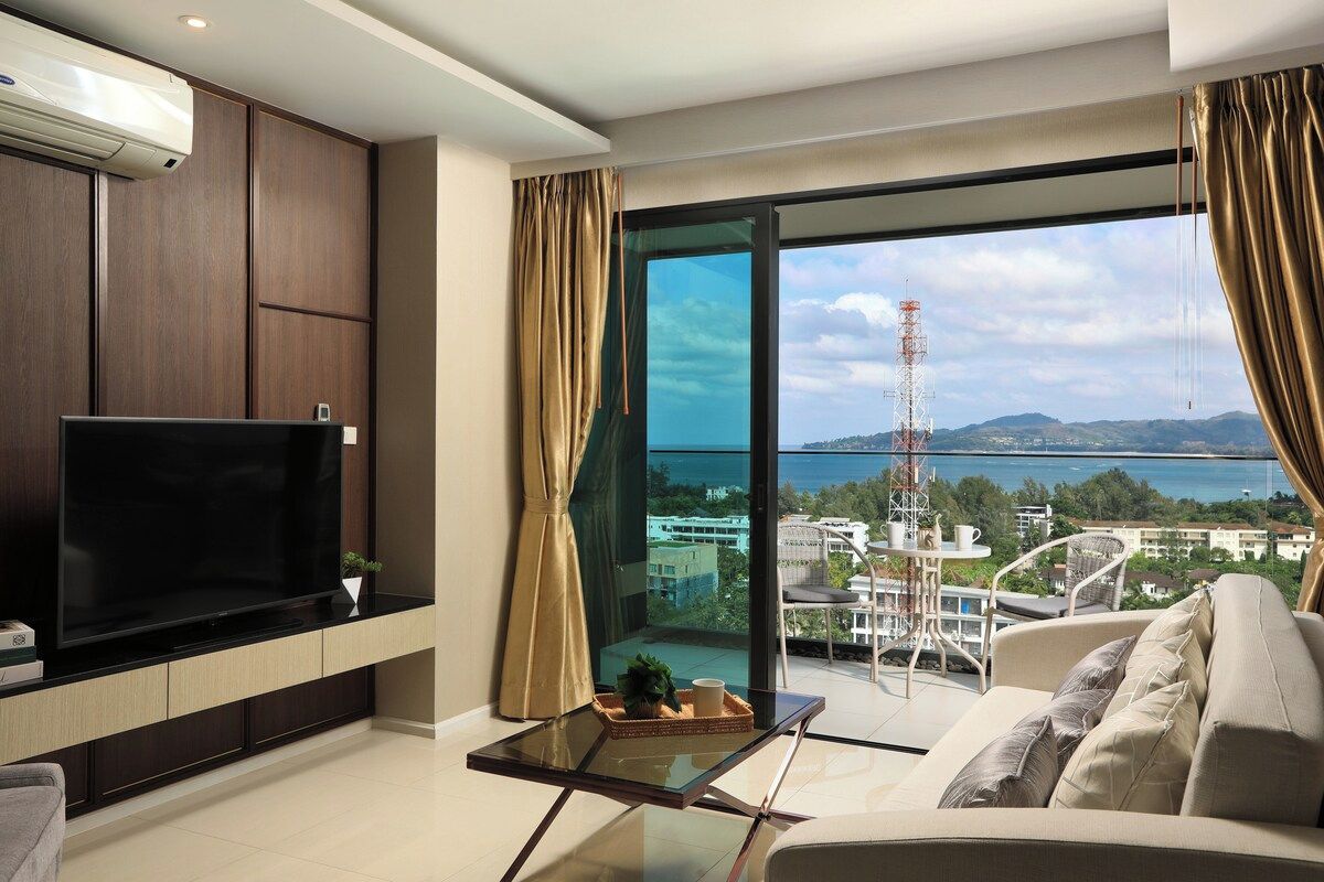 A modern living room with a flat-screen TV, a glass coffee table, and a sofa. A large window offers a view of a cityscape and a body of water.
