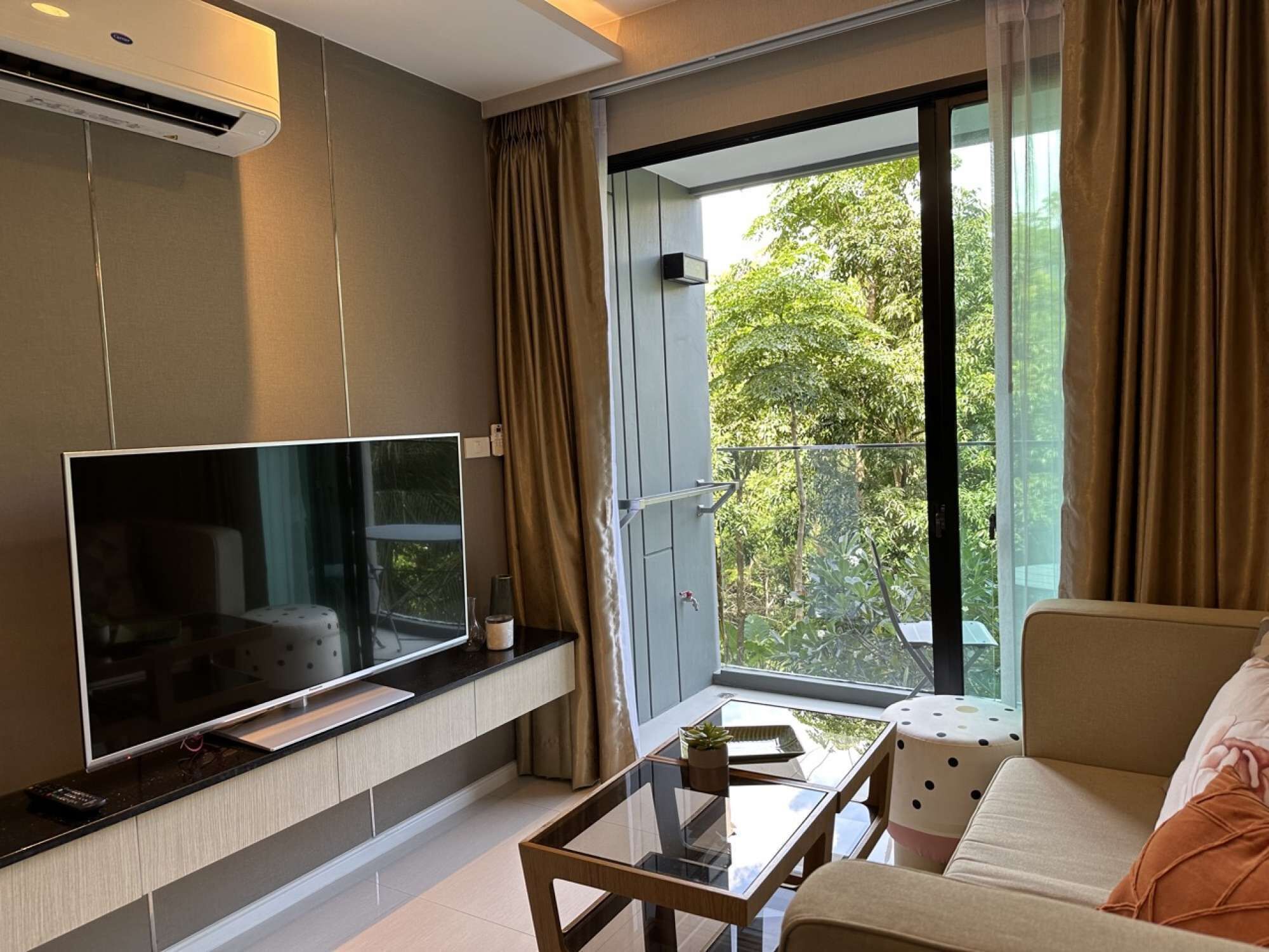 Un salon moderne doté d’un téléviseur à écran plat fixé au mur, d’une table basse en verre et d’un canapé offrant une vue sur la verdure environnante.
