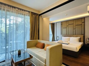 A hotel room with a sofa, a bed, a glass coffee table, and a view of trees through the window. - 1BR Apartment Mountain View | Surin | Capital Pro (Surin)