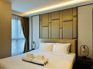 A hotel room with a large bed, a headboard with a padded design, a bedside table with a lamp, and a window with curtains. - 1BR Apartment Mountain View | Surin | Capital Pro (Surin)
