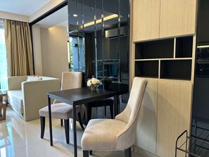 A modern living room with a sofa, a dining table, and chairs. There is a large window with curtains, and a wall-mounted shelf unit. - 1BR Apartment Mountain View | Surin | Capital Pro (Surin)