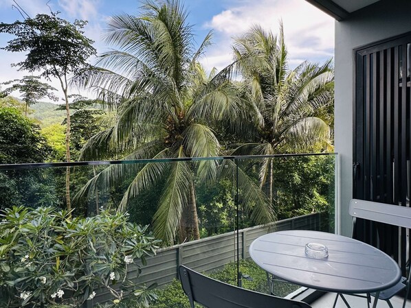 A balcony with a table and chairs, overlooking a lush garden with palm trees. - 1BR Apartment Mountain View | Surin | Capital Pro (Surin)