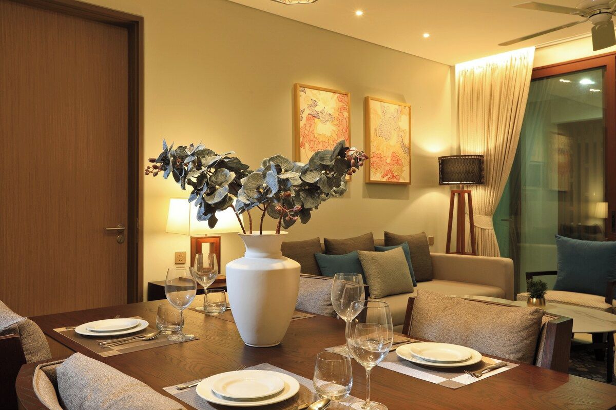 A dining area with a wooden table set for four, a white vase with foliage, and two abstract paintings on the wall.