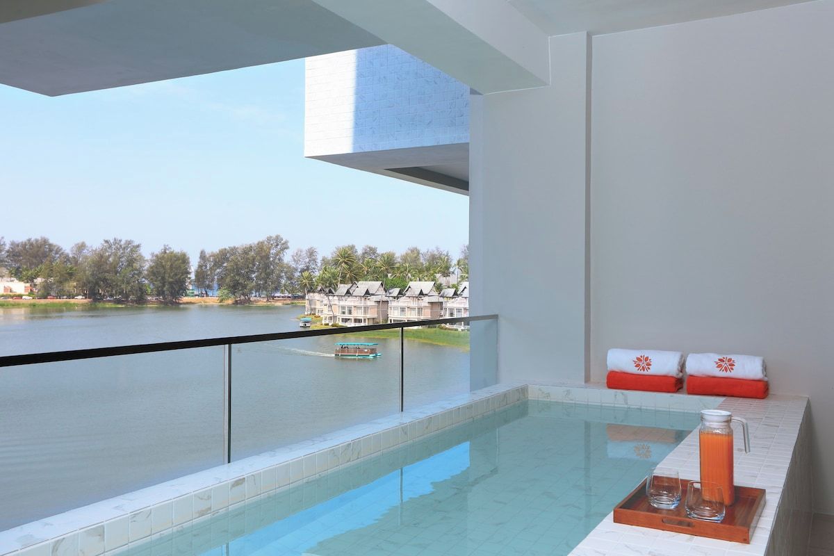 A modern hotel room with a glass balcony overlooking a lake and a row of buildings.