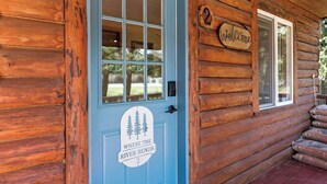 A log cabin with a blue door, a welcome sign, and a window with a view of the outdoors. - Authentic Montana log cabin, 15 min to Glacier. Queen bed + twin bunks, full kitchen & covered porch. Real adventure, real comfort — no tent required. (Martin City)