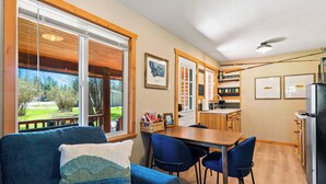A cozy room with a blue sofa, a wooden dining table, and chairs. There is a kitchen area with a refrigerator and a coffee machine. - Authentic Montana log cabin, 15 min to Glacier. Queen bed + twin bunks, full kitchen & covered porch. Real adventure, real comfort — no tent required. (Martin City)