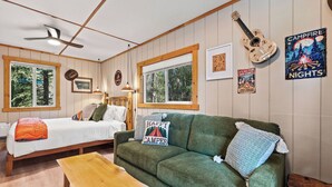 Et hyggeligt værelse i sommerhusstil med seng, grøn sofa, træbord og en guitar på væggen.