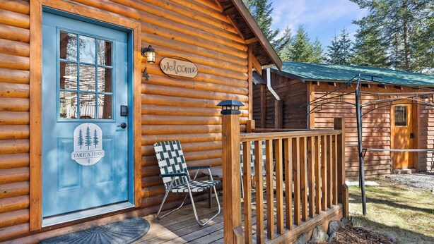Eine Holzhütte mit blauer Tür, einem 'Welcome'-Schild, einem 'Take a Hike'-Schild, einem Stuhl und einer Veranda.