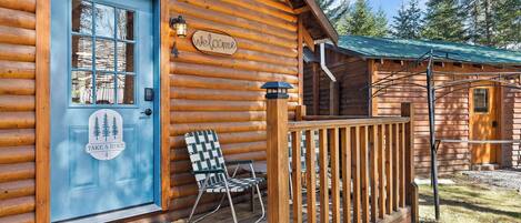 Une cabane en rondins avec une porte bleue, une pancarte « Bienvenue », une pancarte « Partez en randonnée », une chaise et une véranda.