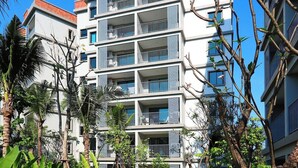 Un hôtel à plusieurs étages doté de balcons, entouré de verdure et bordé d’une promenade dallée.