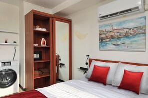 A hotel room with a bed, a wooden wardrobe, a washing machine, a painting, and an air conditioning unit. - Garden View Studio near Maikhao Beach |Capital Pro (Mai Khao)