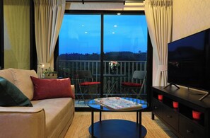 Ein Zimmer mit einer Couch, zwei Sesseln, einem Tisch mit einer Zeitschrift, einem Fernseher und einem Balkon mit Geländer.