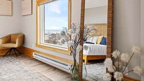 Ein Schlafzimmer mit großem Fenster, einem Bett mit Kissen, einem Sessel und einem Spiegel.