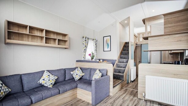 Una sala de estar moderna con un sofá modular gris, estantes de madera, una escalera y una zona de cocina.