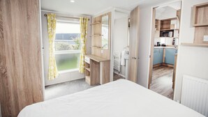 Un dormitorio con una cama, un escritorio, una ventana con cortinas y una puerta abierta que da a una cocina.