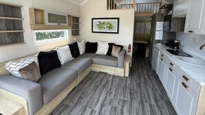 A modern interior with a sectional sofa, wooden shelves, a kitchen with white cabinets, and a hanging light fixture.