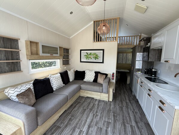 A modern interior with a sectional sofa, wooden shelves, a kitchen with white cabinets, and a hanging light fixture.