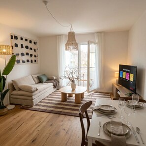 Ein modernes Wohnzimmer mit einer Couch, einem Couchtisch, einem Esstisch für vier Personen und einem Fernseher, der einen Streaming-Dienst zeigt.