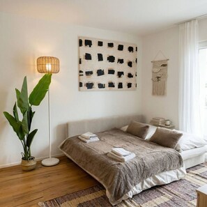 A bedroom with a bed, a floor lamp, a wall art piece, a hanging macrame, and a potted plant. - Riverfront 1BR & Balcony | Zurich Airport | WiFi (Opfikon)