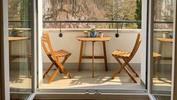 Ein Balkon mit zwei Holzstühlen, einem kleinen Tisch, einer hängenden Lampe und Blick auf Bäume und Gebäude.