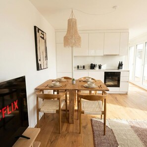 A modern kitchen with white cabinets, a wooden dining table with four chairs, and a hanging light fixture. - Riverfront 1BR & Balcony | Zurich Airport | WiFi (Opfikon)