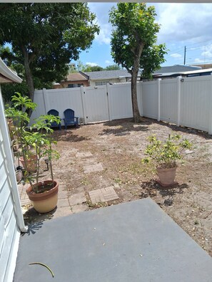 A backyard with a white fence, potted plants, a blue chair, and a tree.